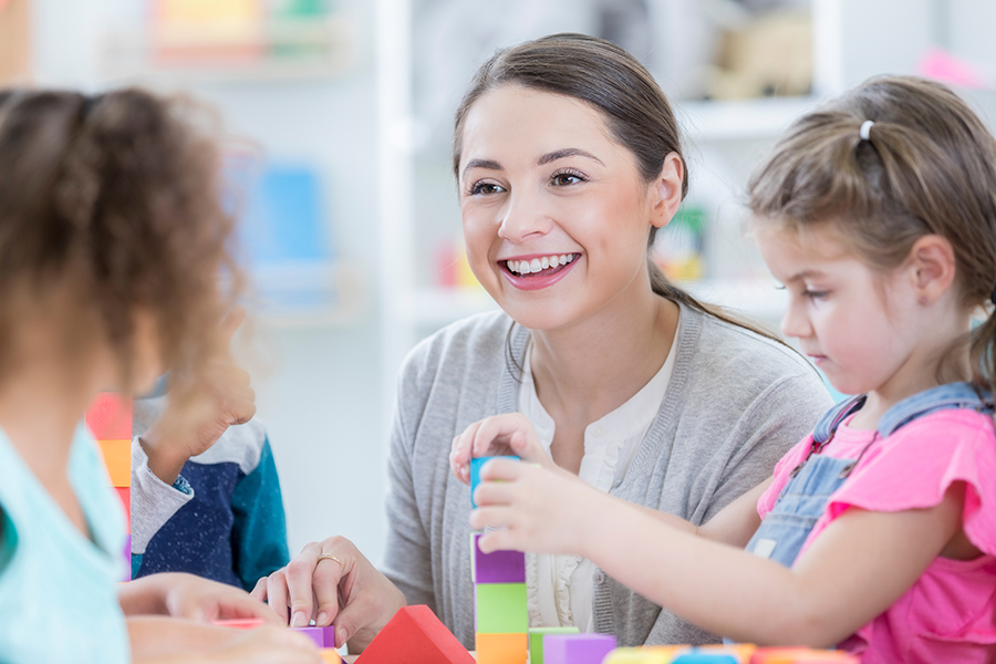 A beautiful young kindergarten teacher sits at a table at school with a few students as they play with multicolored blocks.  She looks across the table and smiles and talks to an unrecognizable student.