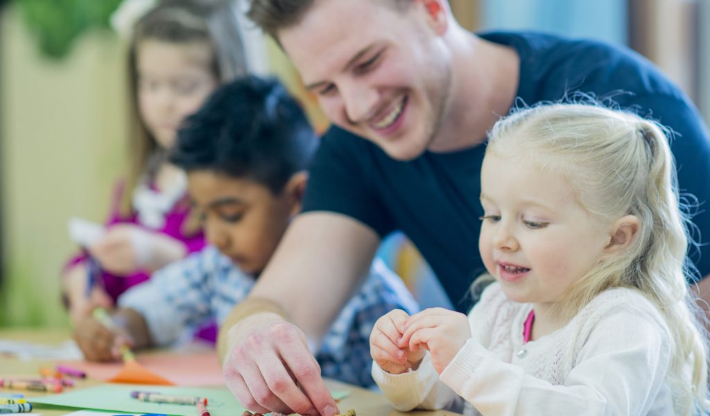 A multi-ethnic group of young children are indoors at a preschool. They are wearing casual clothing. Their male teacher is helping them color with crayons. A Caucasian girl is smiling in front.