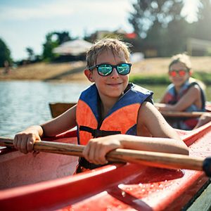boy wearing sunglasses smiling and holding a paddle in a kayak with another child in background
